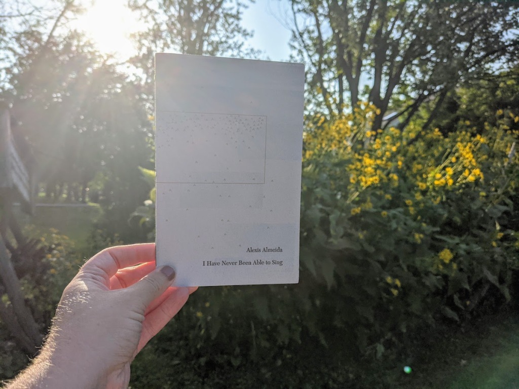 a pale white hand and wrist holds a small flecked book in front of flowers and sun. the book says Alexis Almeida I Have Never Been Able To Sing
