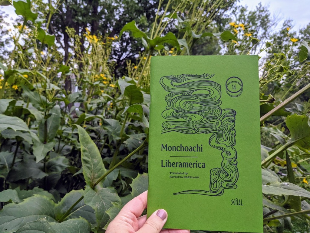 a white hand holds a small green book with orante swirls that might be snakes or gnarled trees: MONCHOACHI'S LIBERAMERICA, translated by Patricia Hartland for Ugly Duckling Presse. in the background are yellow flowers, seed pods, and green foliage almost the same shade as the book.