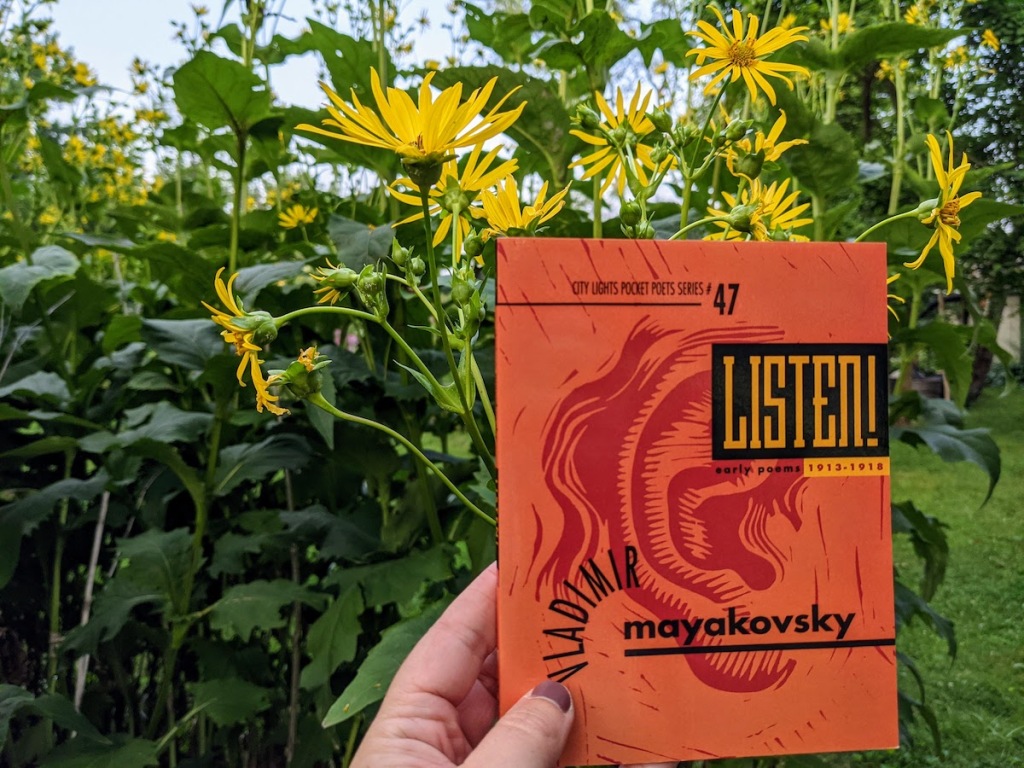 a white hand holds LISTEN! EARLY POEMS 1913-1918 by Vladimir Mayakovsky, a small orange book in front of yellow flowers.